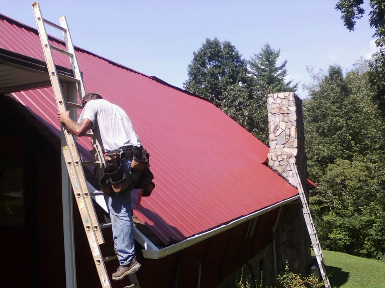 Roofer on a ladder beside a red metal roof and stone chimney — Soffit & Fascia Repair by Roofing Brothers in Belton